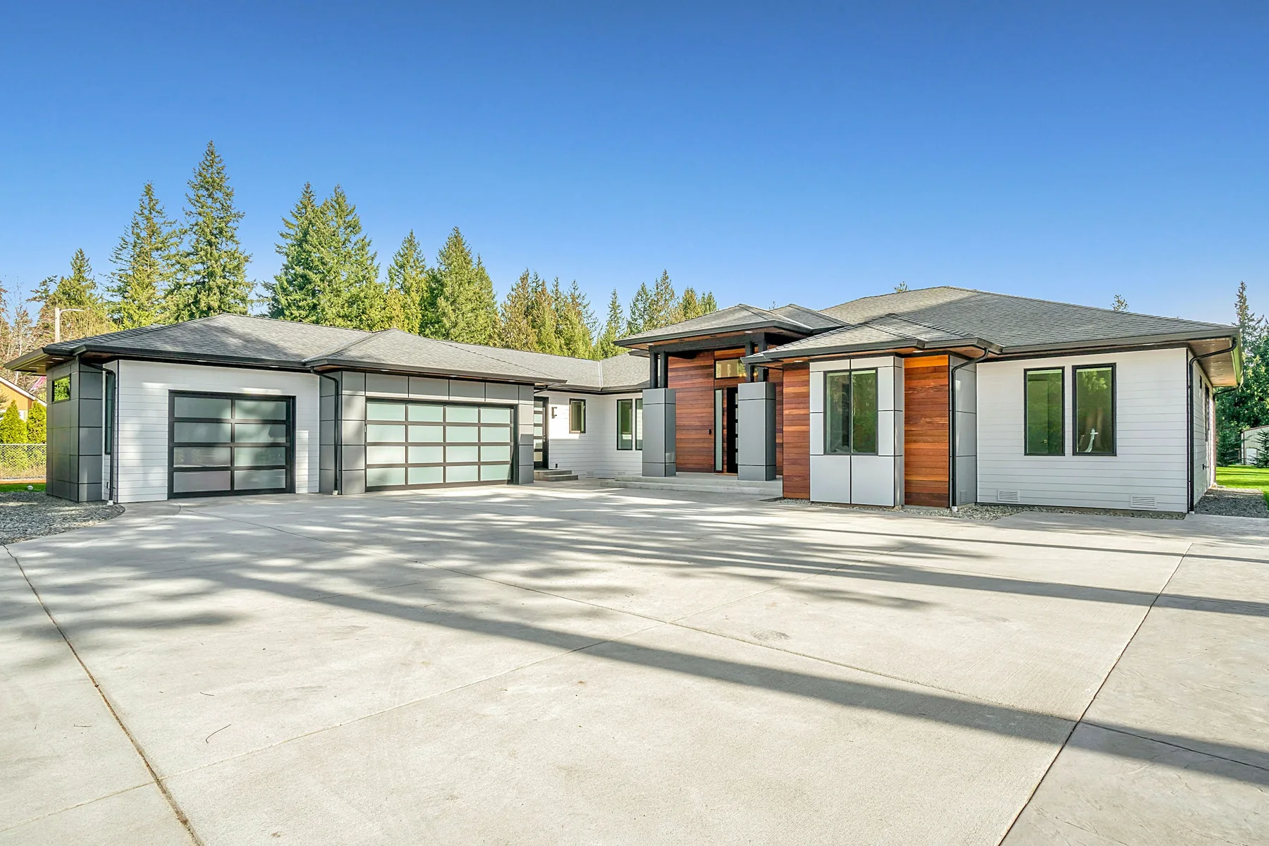 Architectural reference: a single-storey contemporary home with cedar and white cladding, a wide driveway, and evergreen trees under a blue sky