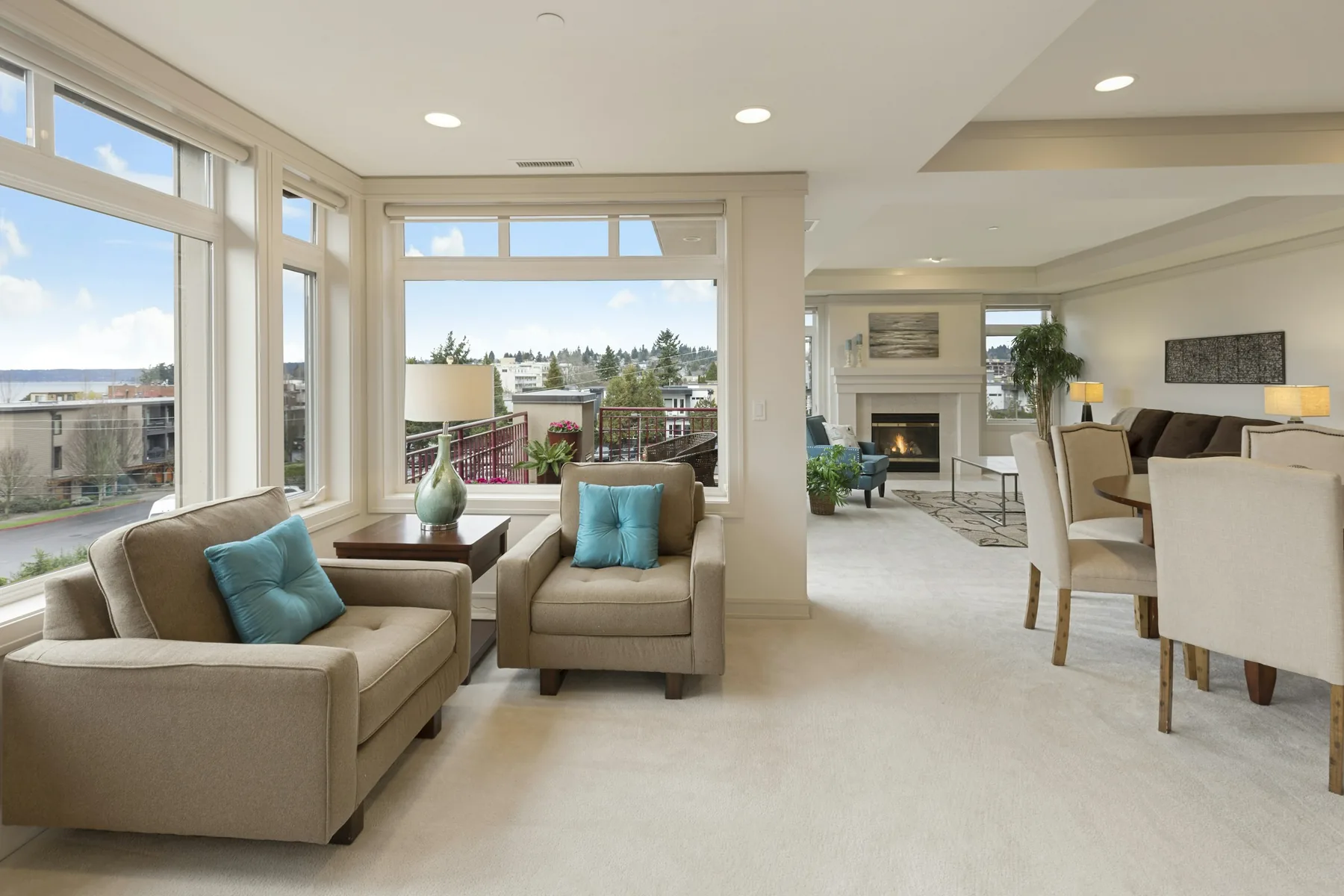 Bright open living room with beige armchairs, large corner windows, a fireplace, and a dining area in a neutral palette